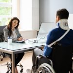 An injured man sitting in a wheelchair across from an official woman at her desk.