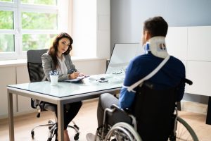 An injured man sitting in a wheelchair across from an official woman at her desk.