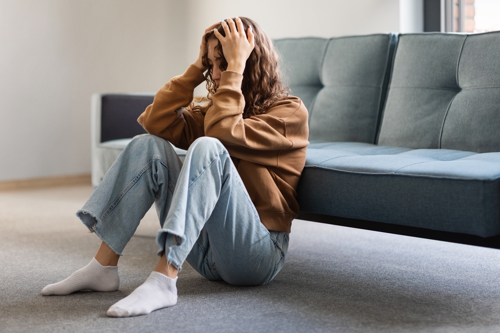Mental Illness Qualify for SSDI - A young woman sitting on the ground with her back against the couch, her elbows propped against her knees as she holds her head in her hands.