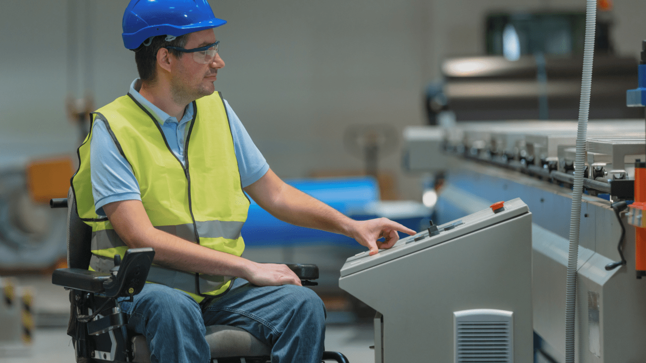 Industrial worker in wheelchair wearing blue hard hat, safety glasses and high-visibility vest operating machinery control panel in modern manufacturing facility Can You Get Both SSI and SSDI