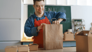 Smiling food service worker in blue shirt and red apron packing a brown paper takeout bag in a restaurant kitchen, with various food containers on the counter