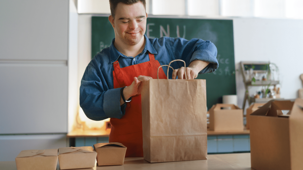 Smiling food service worker in blue shirt and red apron packing a brown paper takeout bag in a restaurant kitchen, with various food containers on the counter
