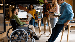 A diverse group of five colleagues in a modern office having a collaborative business meeting, with one team member in a wheelchair presenting to the group near a monitor displaying quarterly report data