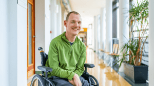 Person in wheelchair wearing green hoodie smiling in modern office hallway