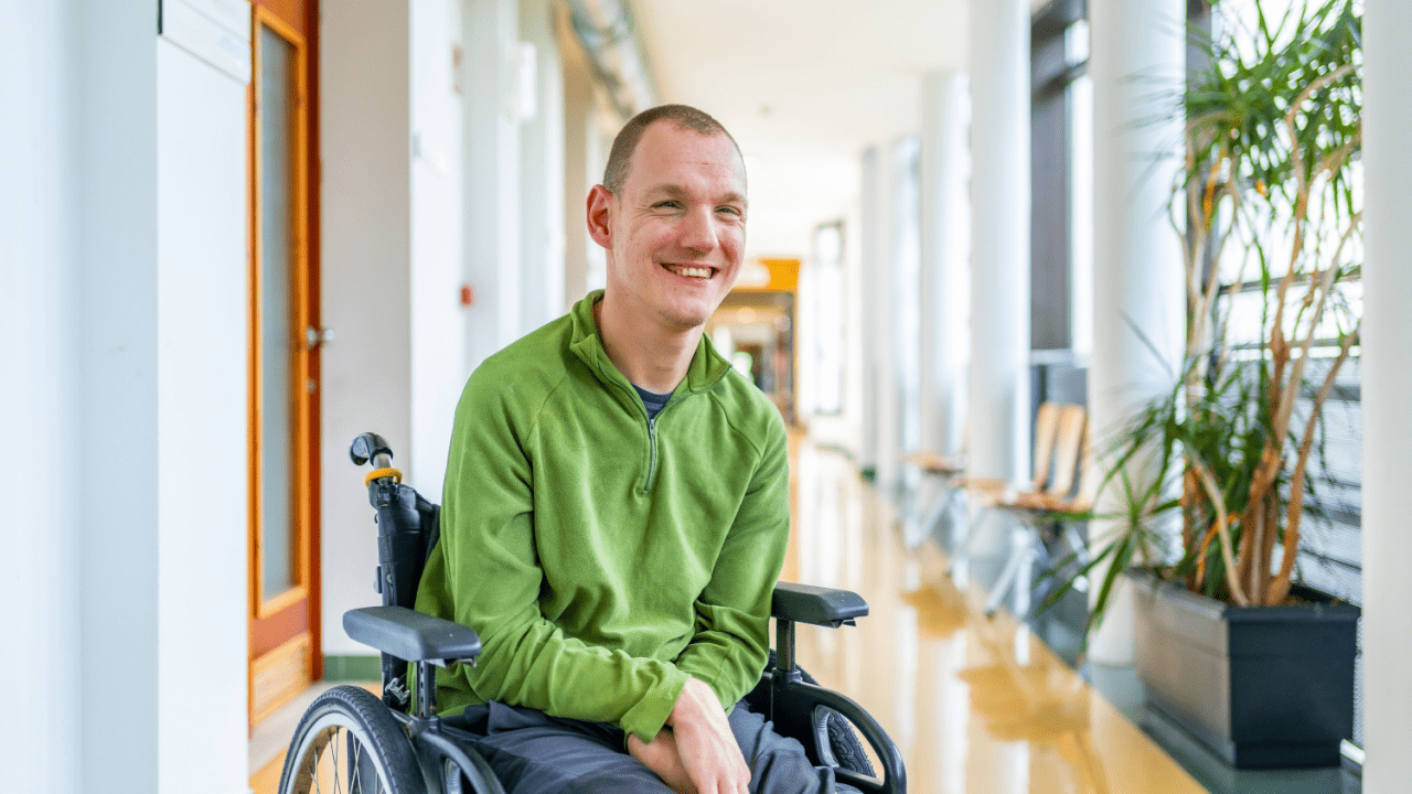 Person in wheelchair wearing green hoodie smiling in modern office hallway