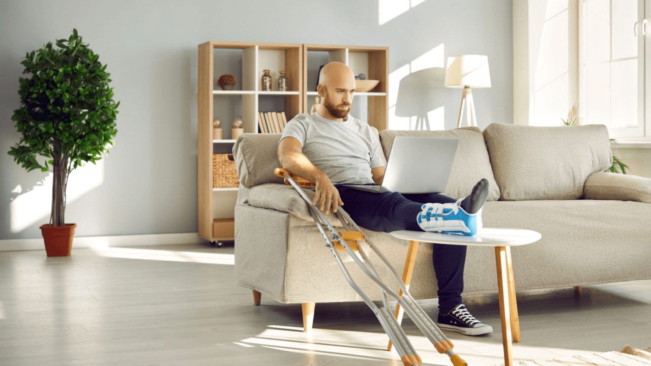 Man with leg injury in medical boot working on laptop while sitting on couch with crutches nearby in modern living room