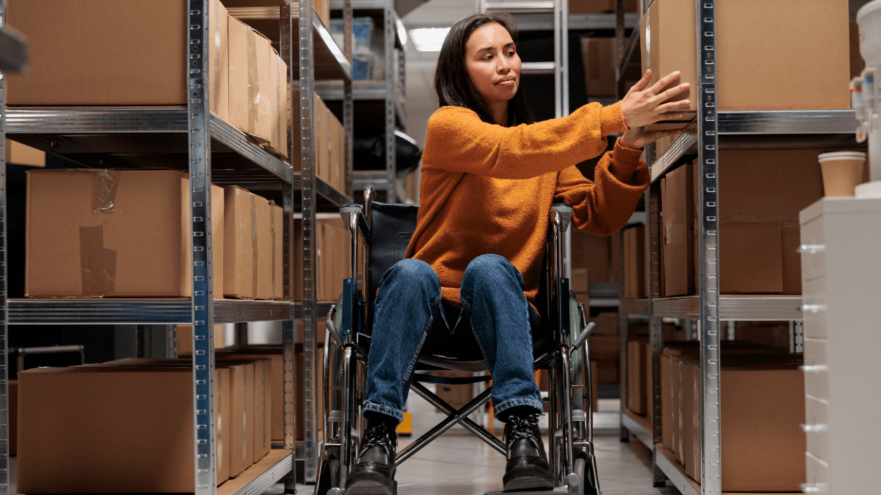 Woman in wheelchair reaching for items on warehouse shelves in accessible workplace environment