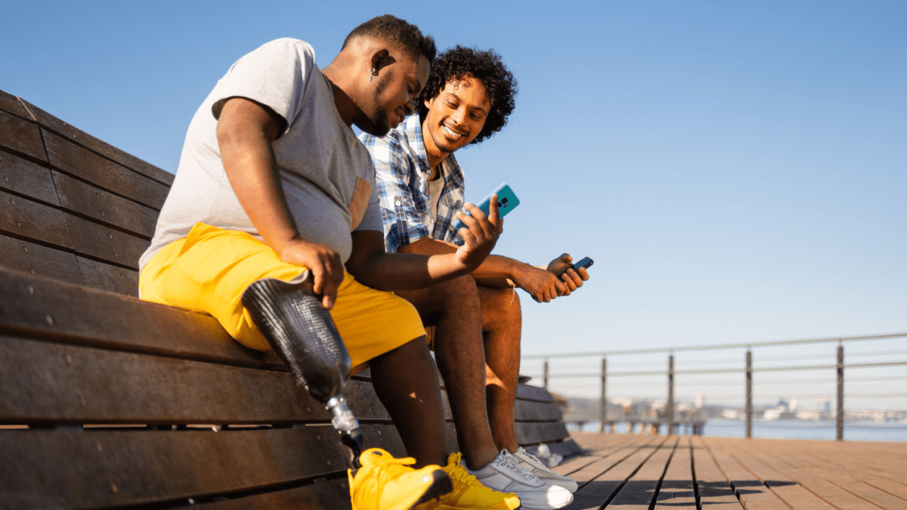 Two young men sitting on a wooden bench outdoors, both looking at their mobile phones, with a clear blue sky and waterfront cityscape in the background