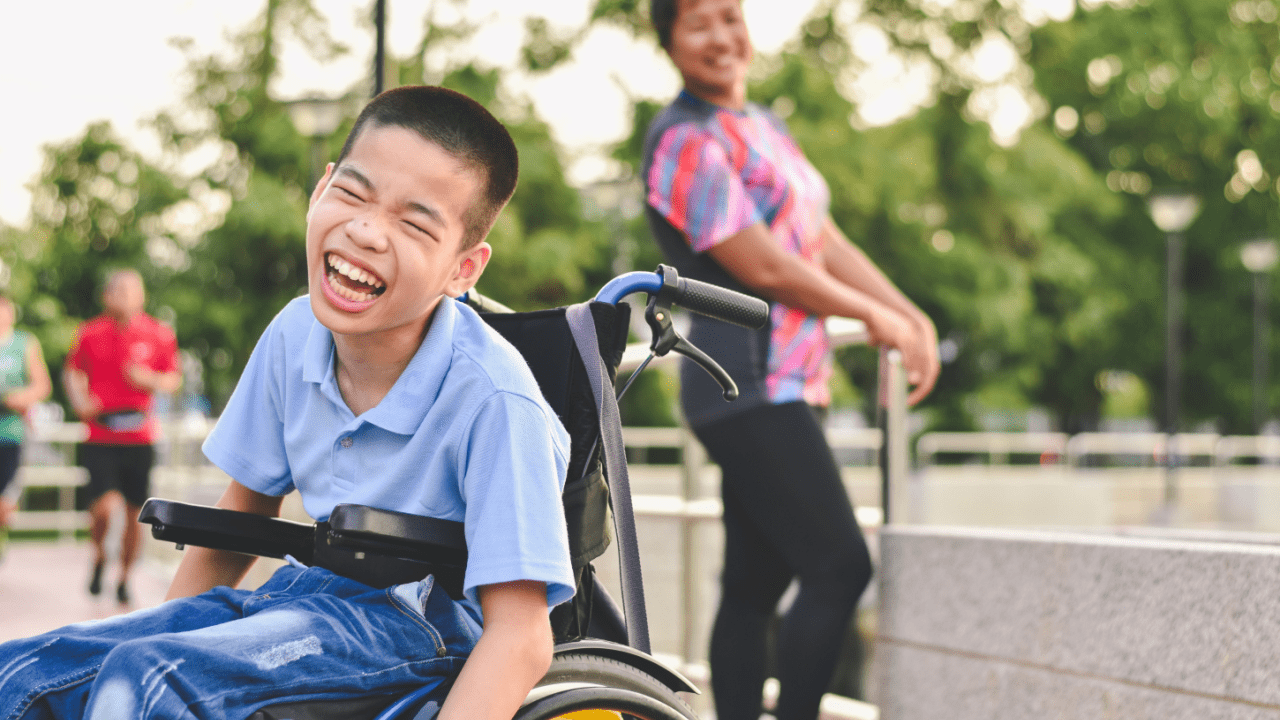 How hard is it to get SSDI Young boy in wheelchair laughing joyfully outdoors in park setting with person in background
