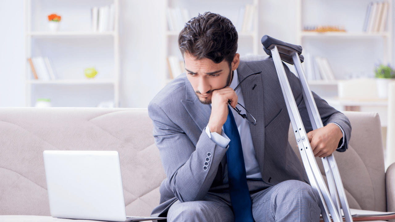Businessman in gray suit looking contemplative while working from home with crutches nearby and laptop on coffee table