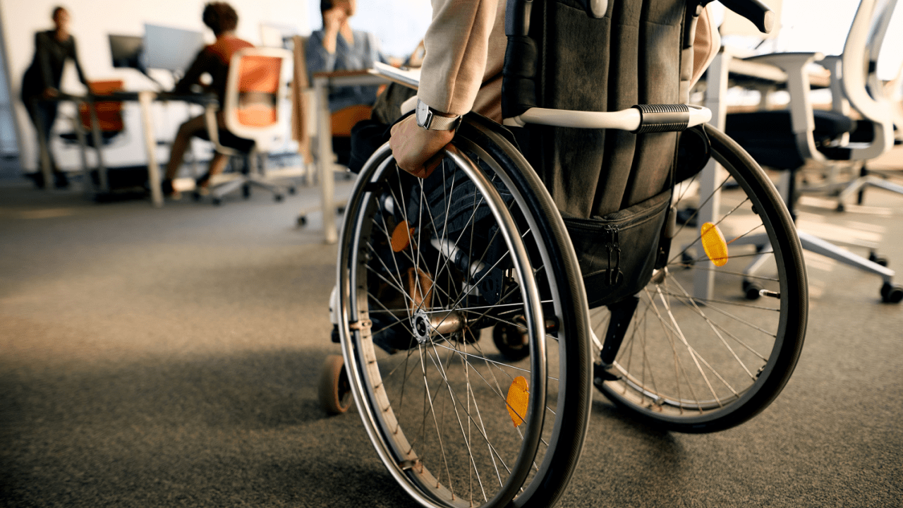 Person in wheelchair at office workspace with colleagues in background