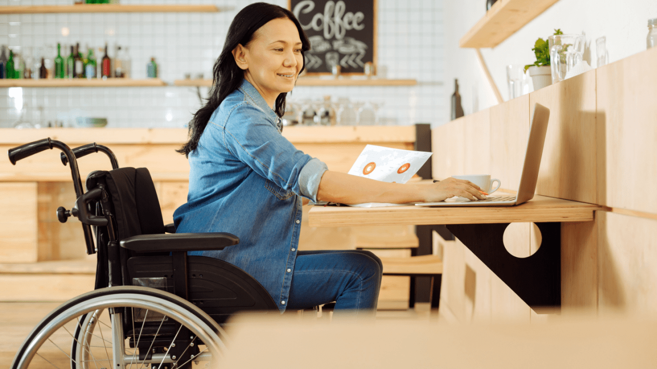 Woman in wheelchair working on laptop at wooden desk in modern kitchen setting