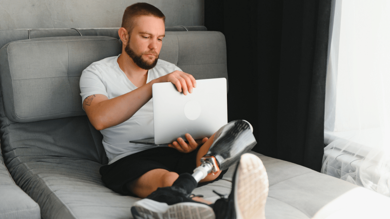 Man with prosthetic leg working on laptop while sitting comfortably on gray sectional sofa