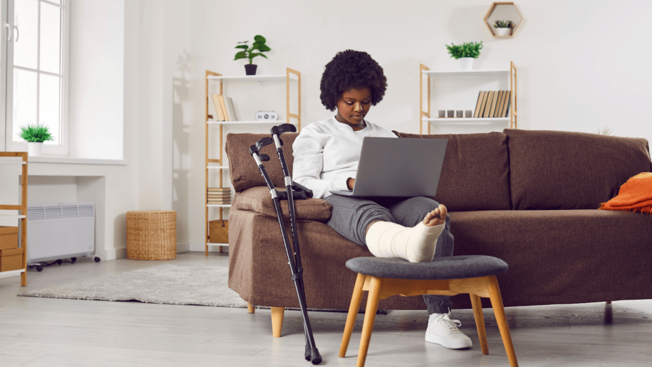 Black woman with foot cast working on laptop from couch with crutches nearby in modern home office