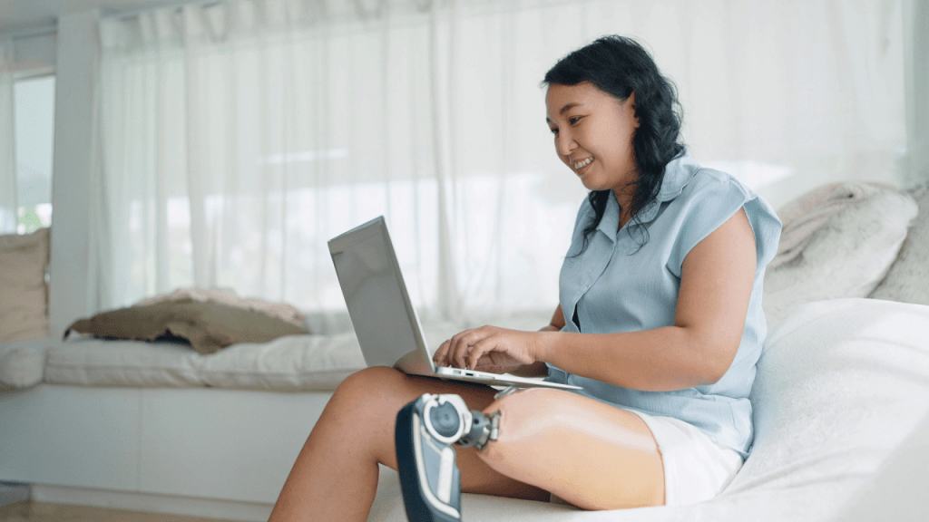 Asian woman with prosthetic leg working happily on laptop while sitting on white couch in bright living room