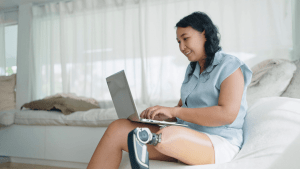 Asian woman with prosthetic leg working happily on laptop while sitting on white couch in bright living room