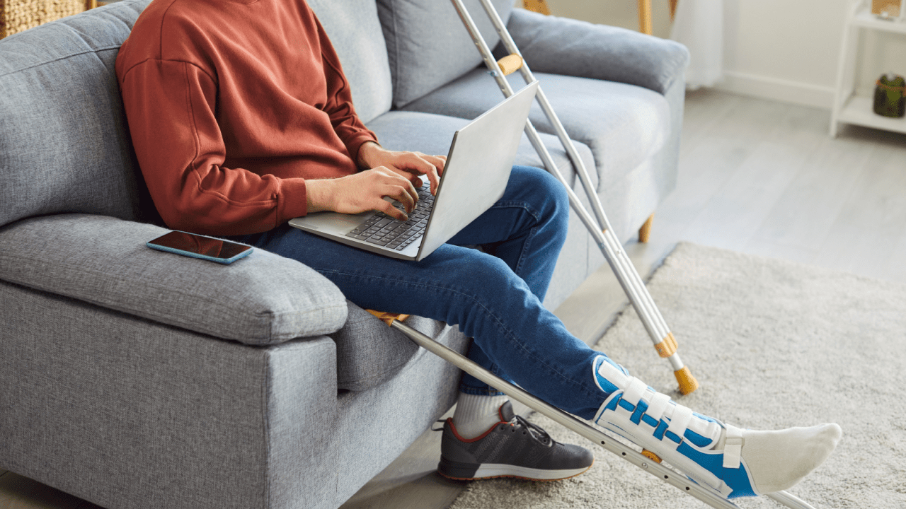 Why do most people get denied for SSDI Person with leg brace working on laptop while sitting on couch with crutches nearby in modern living room