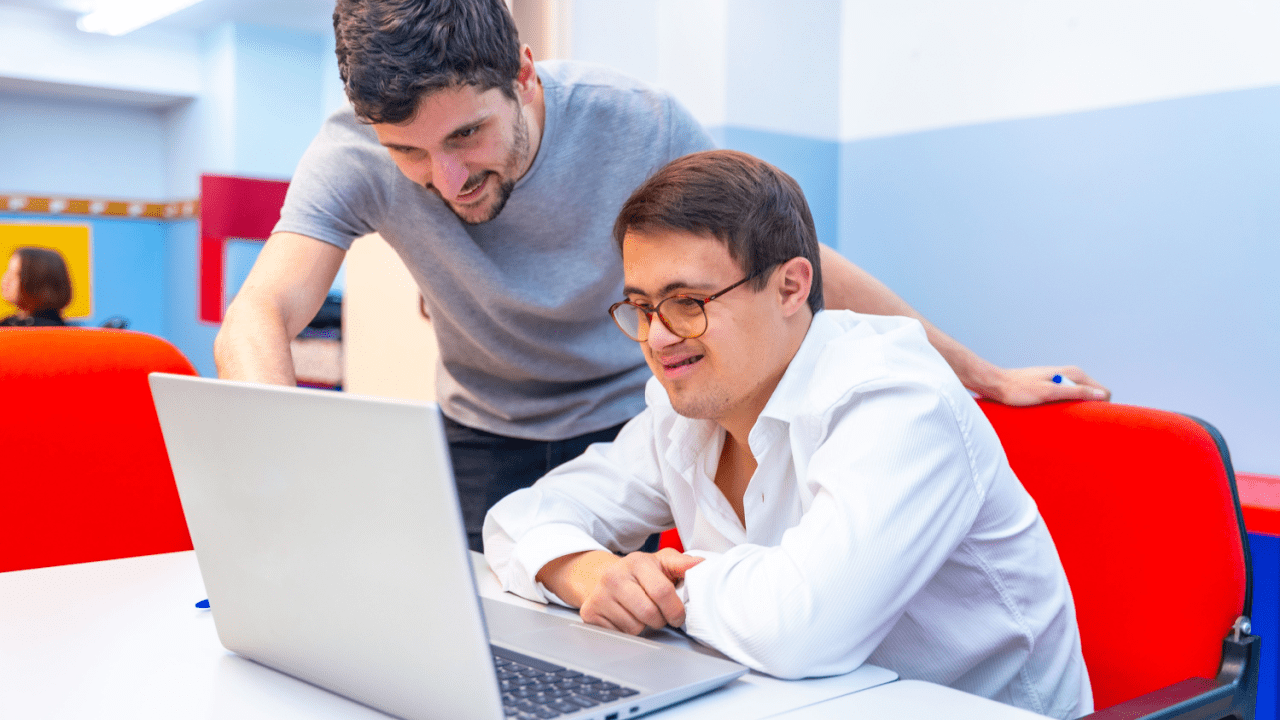 Two professional men collaborating on laptop computer researching what is 100% social security disability pay in modern colorful office workspace
