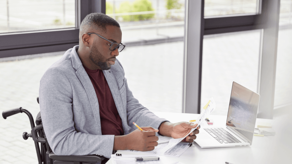 A man in a wheelchair reviewing SSDI documents at a desk while working on a laptop, preparing for a continuing disability review