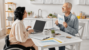 A woman using a wheelchair works on a laptop at a kitchen table while a man reviews printed charts and documents, discussing what are red flags on a disability update report during an SSDI consultation at home.